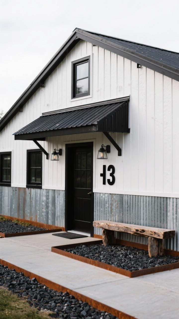 Wide, slightly elevated exterior shot of a high-contrast small barn: upper half in matte white vertical siding, lower half in corrugated galvanized steel; black window frames and matching black metal roof; a bold industrial awning over the entry; warehouse-style bulkhead fixtures beside oversized black house numbers mounted on a metal panel; corten steel edging containing dark basalt gravel paths; a reclaimed timber beam used as a bench at the threshold; crisp, industrial, resilient mood.